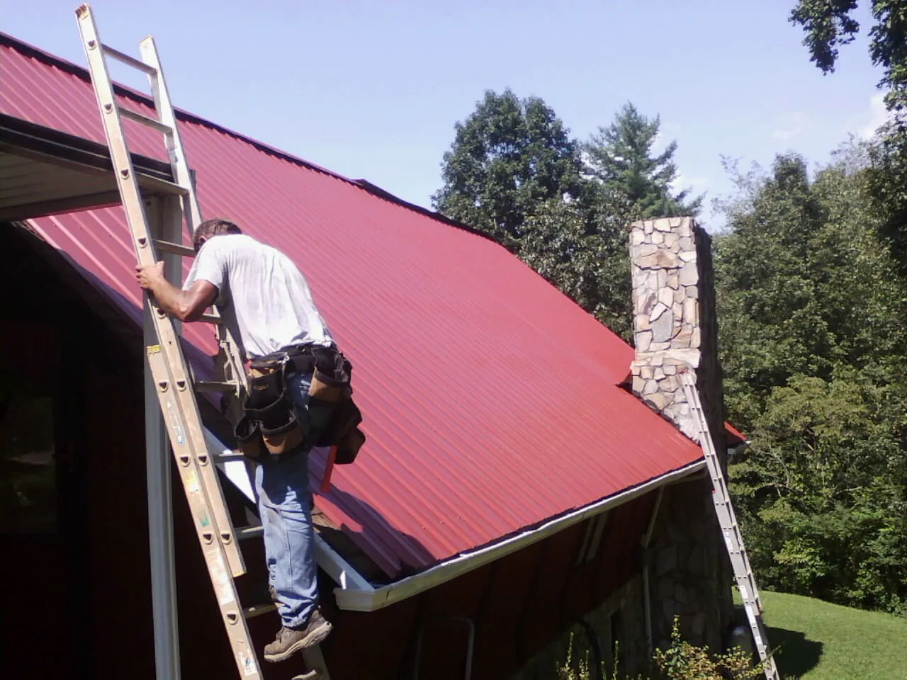 Roofer on a ladder beside a red metal roof and stone chimney — Soffit & Fascia Repair by Roofing Brothers in Plush